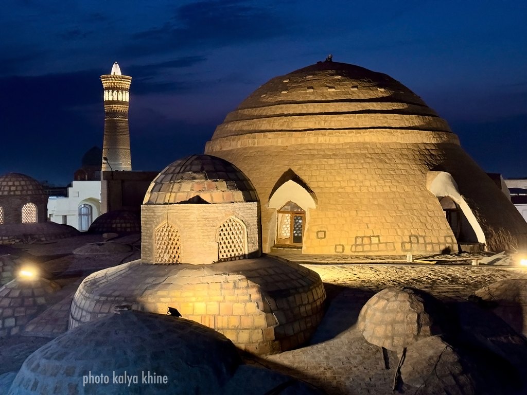 Kuppeln der Altstadt von Bukhara in der blauen Stunde, dahinter ein beleuchtetes Minarett, Usbekistan